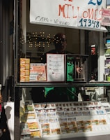 A child holding an ice cream cone stands outside a small kiosk or lottery ticket booth. The booth displays various colorful lottery tickets in the window. There are signs advertising a significant lottery prize amount in Spanish. A person can be seen partially obscured inside the booth, and plants are visible around the area, suggesting an outdoor setting.