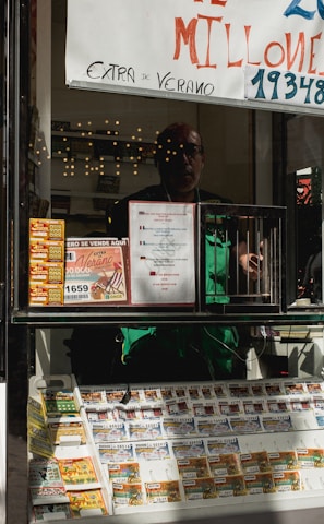A child holding an ice cream cone stands outside a small kiosk or lottery ticket booth. The booth displays various colorful lottery tickets in the window. There are signs advertising a significant lottery prize amount in Spanish. A person can be seen partially obscured inside the booth, and plants are visible around the area, suggesting an outdoor setting.