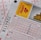 A close-up image of lottery tickets featuring crossed-out numbers, brand logos, and various numbers in a grid format. The tickets have sections for different games, and numbers are marked with black crosses.
