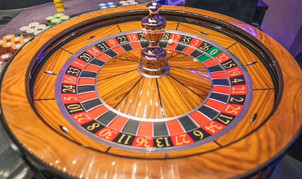 A roulette wheel with alternating red and black numbered pockets on a wooden platform. The wheel contains a small white ball resting in one of the pockets, indicating gameplay. Surrounding the wheel are stacks of colorful poker chips, suggesting a casino environment.
