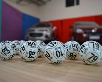 A collection of white lottery balls with black numerals scattered on a wooden surface. Two vehicles are blurred in the background, parked in a room with multicolored walls including teal, purple, blue, and red panels.