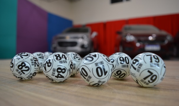 A collection of white lottery balls with black numerals scattered on a wooden surface. Two vehicles are blurred in the background, parked in a room with multicolored walls including teal, purple, blue, and red panels.
