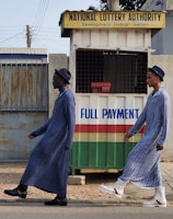 Two individuals are walking past a small kiosk with signage for the National Lottery Authority. The kiosk, decorated with a sign that reads 'Full Payment,' features yellow, green, red, and white colors. The individuals are dressed in matching long blue robes and hats, with one wearing black boots and the other white boots.