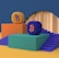 Two platform structures with stairs are positioned side by side. One platform is orange with an orange and purple 'Tezos' symbol on top, while the other is green with a blue and orange 'Bitcoin' symbol. The platforms are set on a blue and mustard yellow background, and there is a large circular arch in the backdrop.