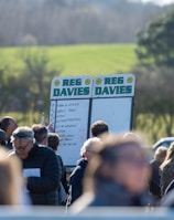 A group of people gathered outdoors with two large betting boards in the background labeled 'REG DAVIES'. The setting appears to be a countryside location with trees and fields visible in the distance. The crowd consists of individuals dressed in jackets and hats, engaging in conversation.