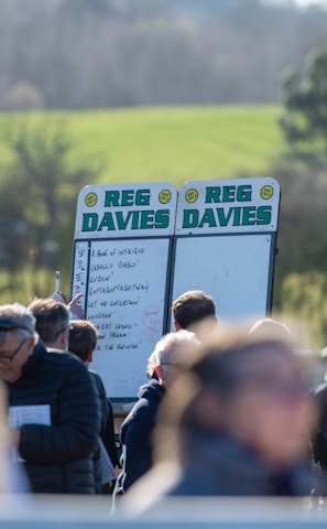 A group of people gathered outdoors with two large betting boards in the background labeled 'REG DAVIES'. The setting appears to be a countryside location with trees and fields visible in the distance. The crowd consists of individuals dressed in jackets and hats, engaging in conversation.
