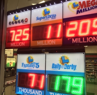 A signboard inside a convenience store displays the current jackpot amounts for various lottery games. There are bright, illuminated numbers in red and green showing the prize amounts: Powerball is at 725 million, Super Lotto Plus at 11 million, Mega Millions at 205 million, Fantasy 5 at 71 thousand, and Daily Derby at 179. In the background, shelves with various drinks and products are visible, along with promotional posters.