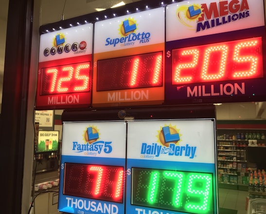 A signboard inside a convenience store displays the current jackpot amounts for various lottery games. There are bright, illuminated numbers in red and green showing the prize amounts: Powerball is at 725 million, Super Lotto Plus at 11 million, Mega Millions at 205 million, Fantasy 5 at 71 thousand, and Daily Derby at 179. In the background, shelves with various drinks and products are visible, along with promotional posters.