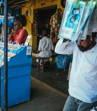 A street vendor stall displaying an array of lottery tickets clipped to a bright blue kiosk. A man in a red shirt leans on the counter, appearing thoughtful, while another man in a white shirt adjusts his hat and walks by. A variety of products hangs from strings above, and a colorful display fills the background with vibrant signs and packets. The scene suggests a busy, lively marketplace.