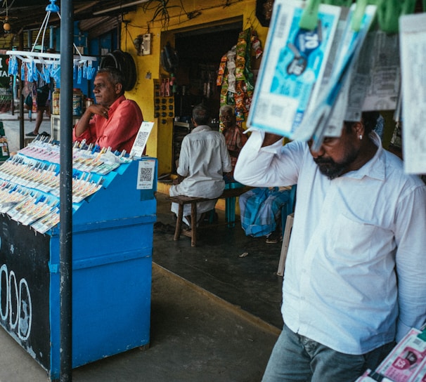 A street vendor stall displaying an array of lottery tickets clipped to a bright blue kiosk. A man in a red shirt leans on the counter, appearing thoughtful, while another man in a white shirt adjusts his hat and walks by. A variety of products hangs from strings above, and a colorful display fills the background with vibrant signs and packets. The scene suggests a busy, lively marketplace.